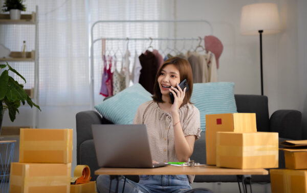 Smiling happy young Asian woman entrepreneur receiving phone cal Smiling happy young Asian woman entrepreneur receiving phone call for new sales order among boxes of product with laptop computer. Success online business and eCommerce concept.